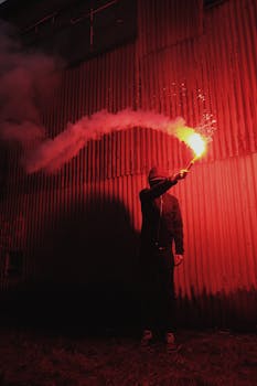 夜间，一个人在波纹金属墙前举着明亮的照明弹，投射出戏剧性的阴影