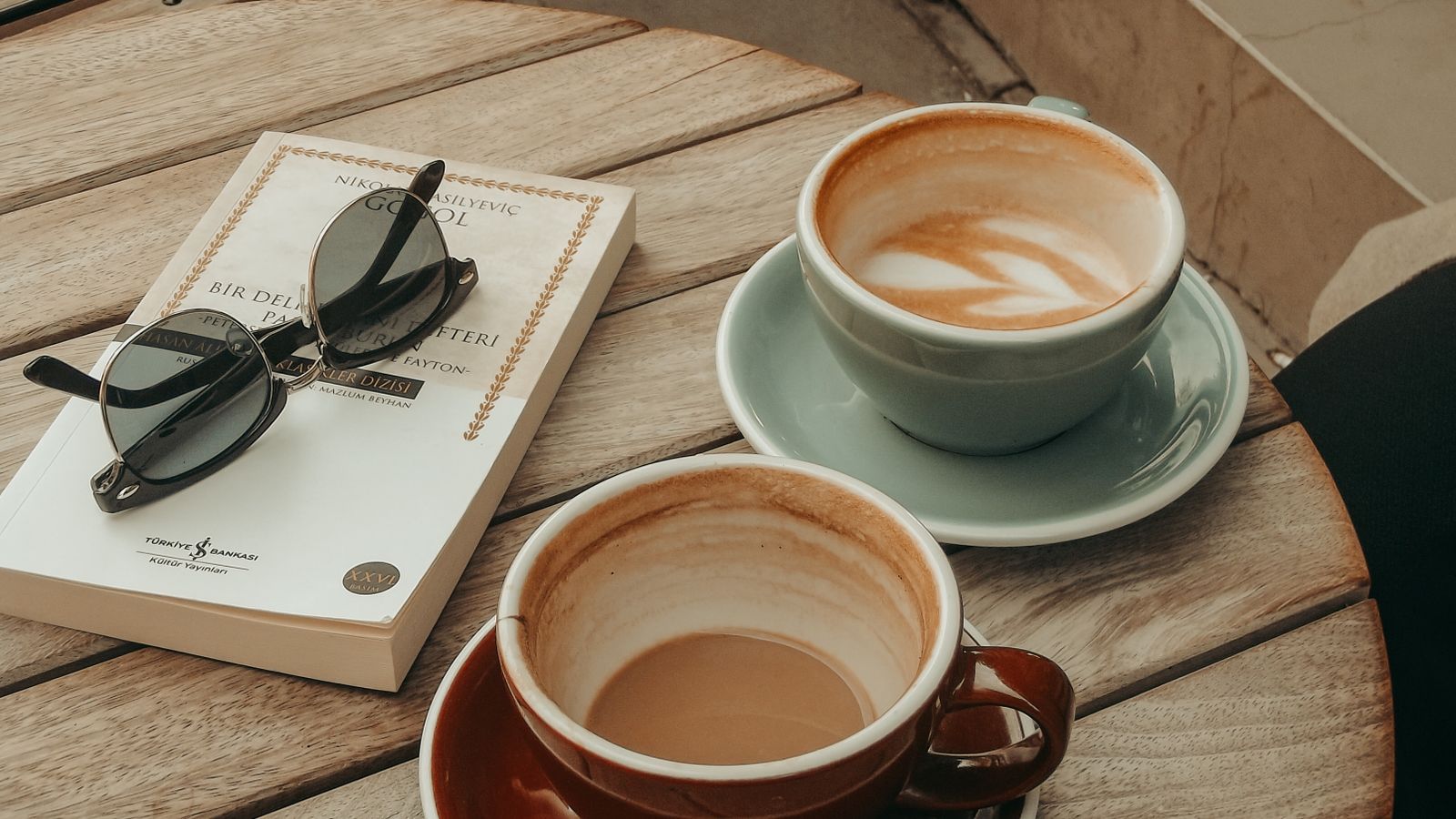 Two cups of coffee beside a book and sunglasses on a wooden café table