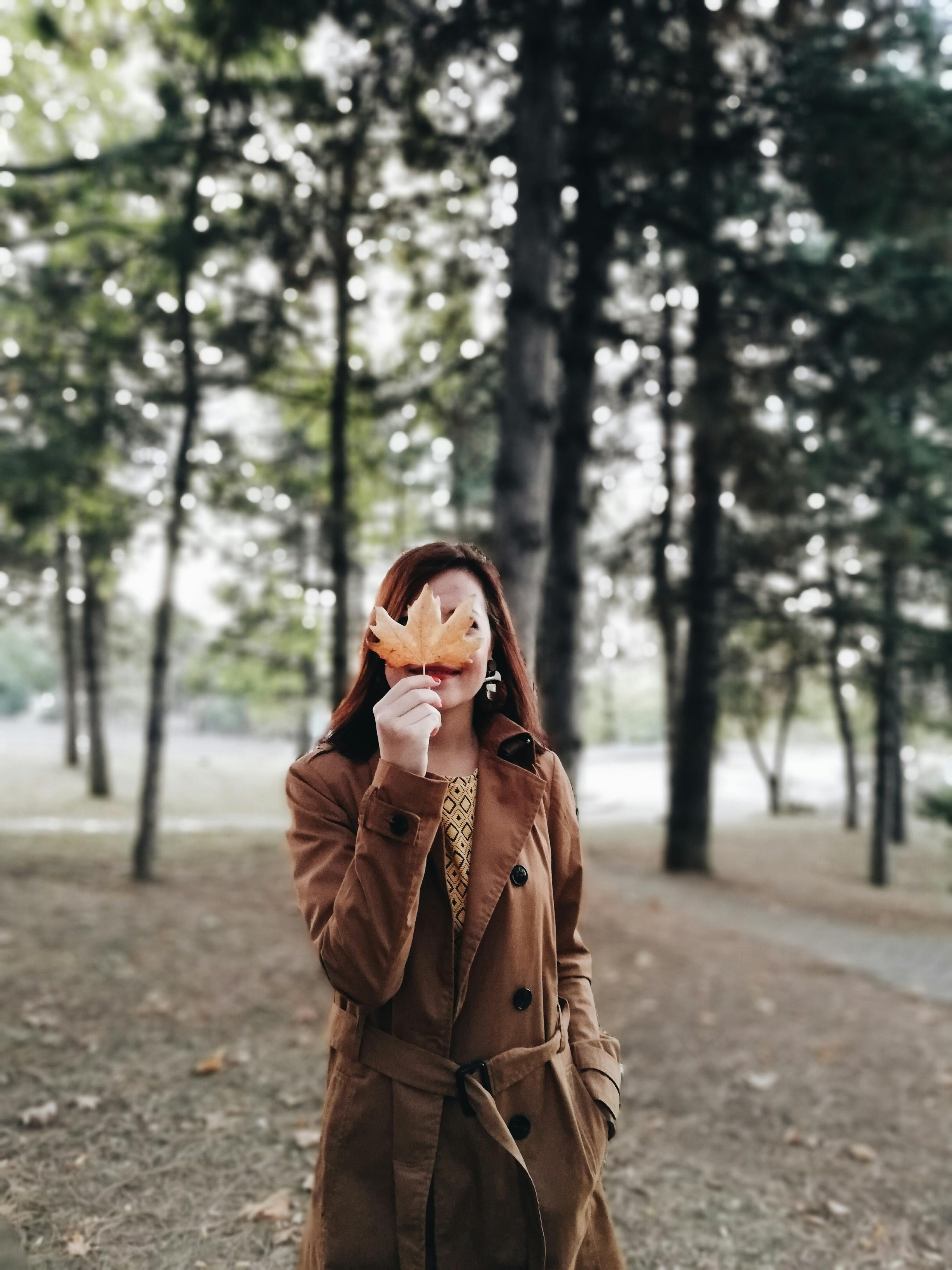 Woman in a brown coat standing in a forest holding a maple leaf, capturing the essence of fall.