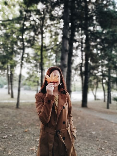 Woman in a brown coat standing in a forest holding a maple leaf, capturing the essence of fall.