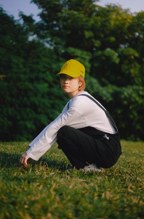 Free A young woman wearing a vibrant cap and stylish outfit crouches on grass outdoors. Stock Photo
