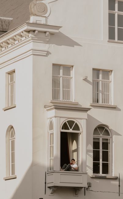 Man relaxing on window ledge of a classic white building exterior, showcasing architectural details.