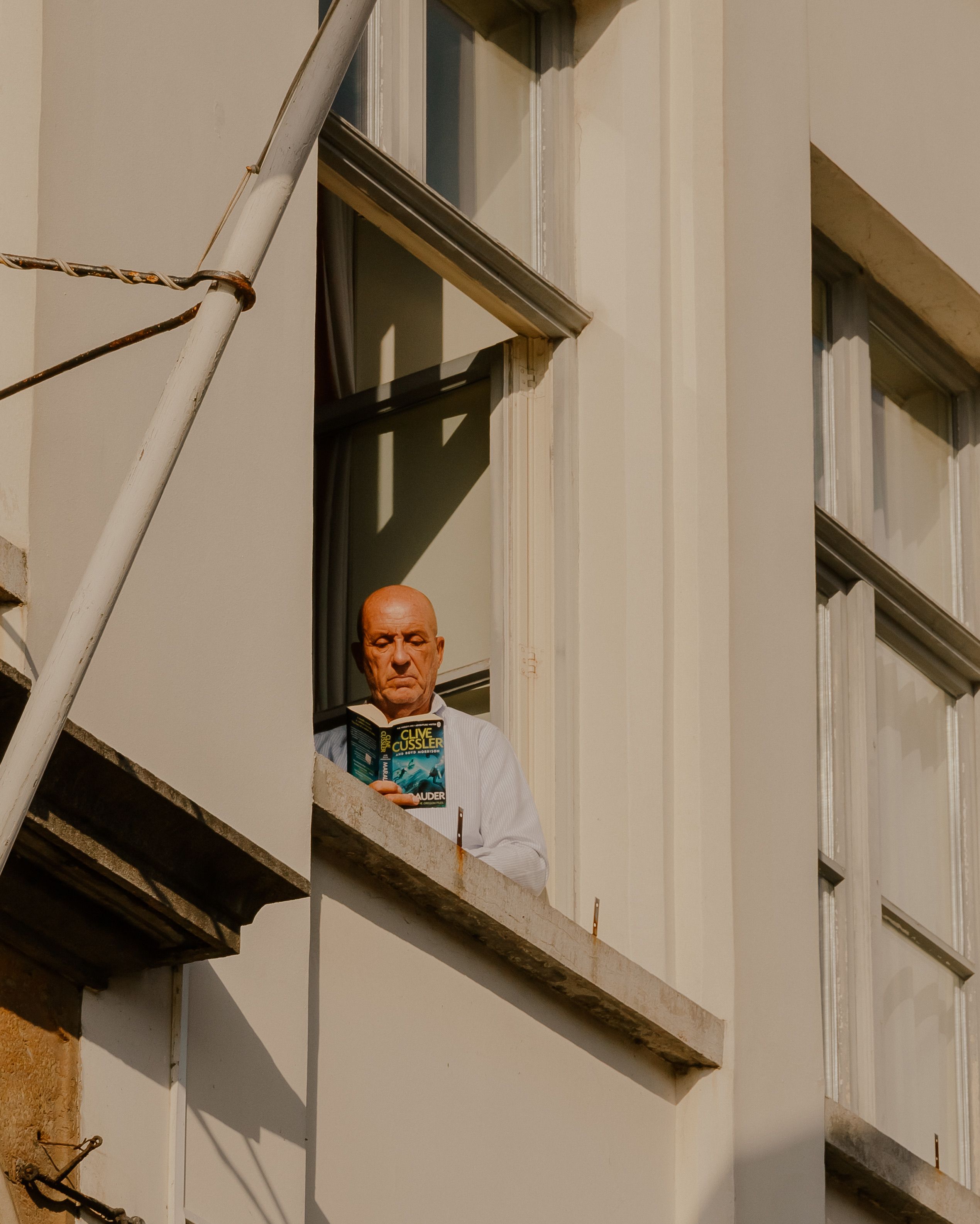 A bald man enjoying a book while standing by a sunny window.