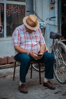 Adult man wearing a hat sitting outdoors on a chair using a smartphone next to a bicycle, displaying modern lifestyle.