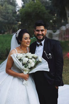 Bride and groom smiling outdoors in elegant wedding attire holding bouquet.