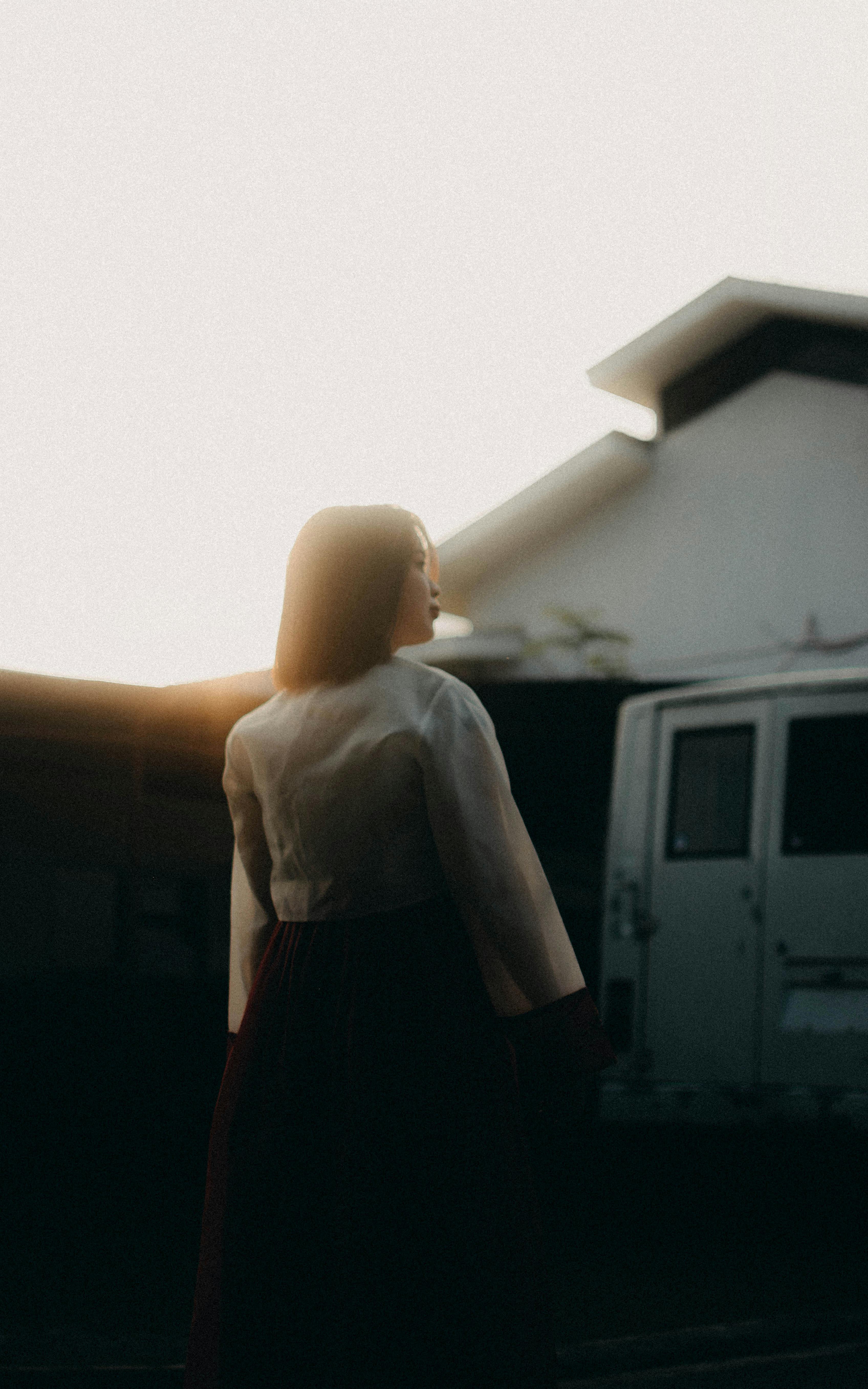 A woman in traditional attire stands silhouetted in front of a house and trailer during sunset.