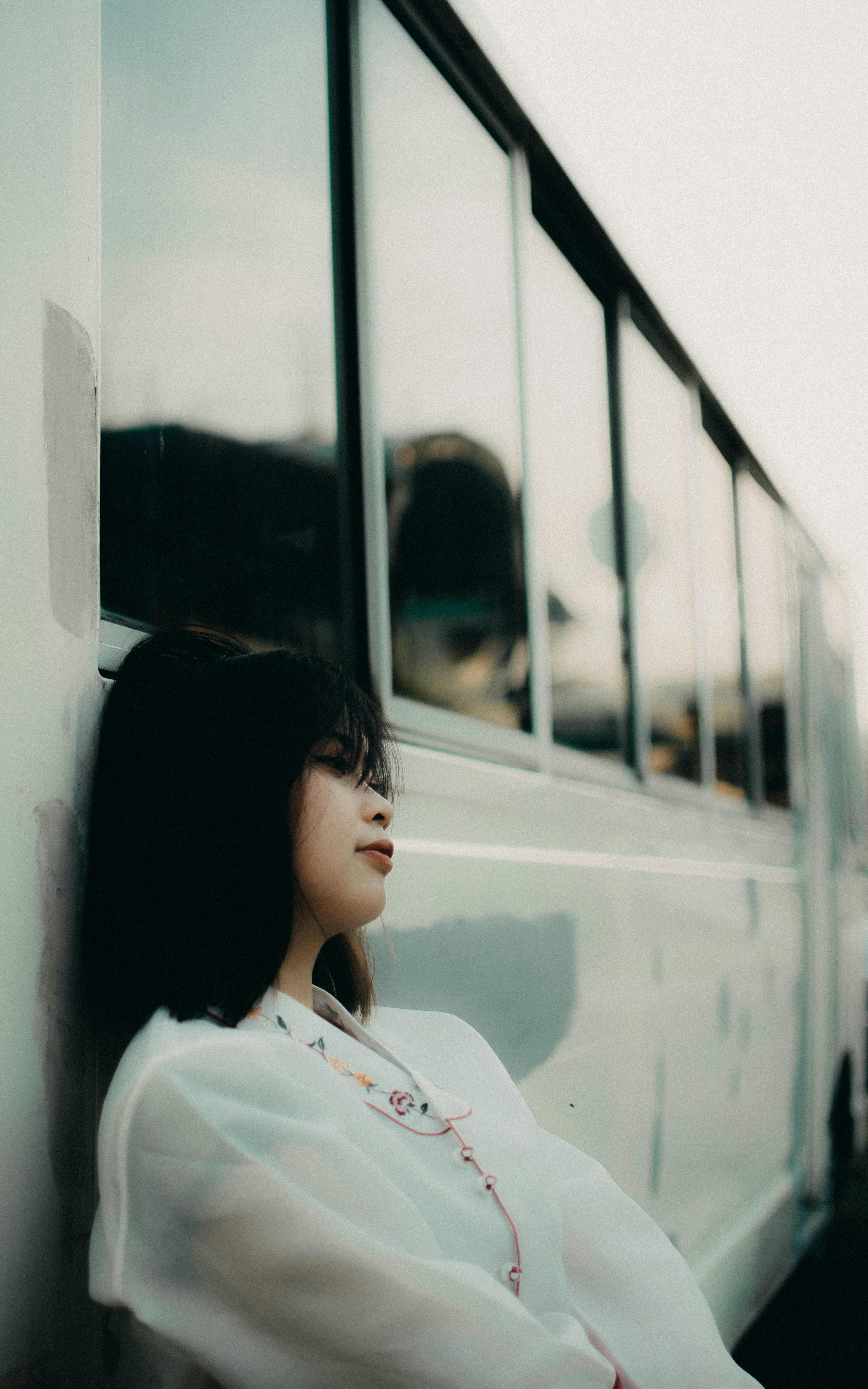 Young woman in white blouse leaning against a bus, lost in thought outdoors.