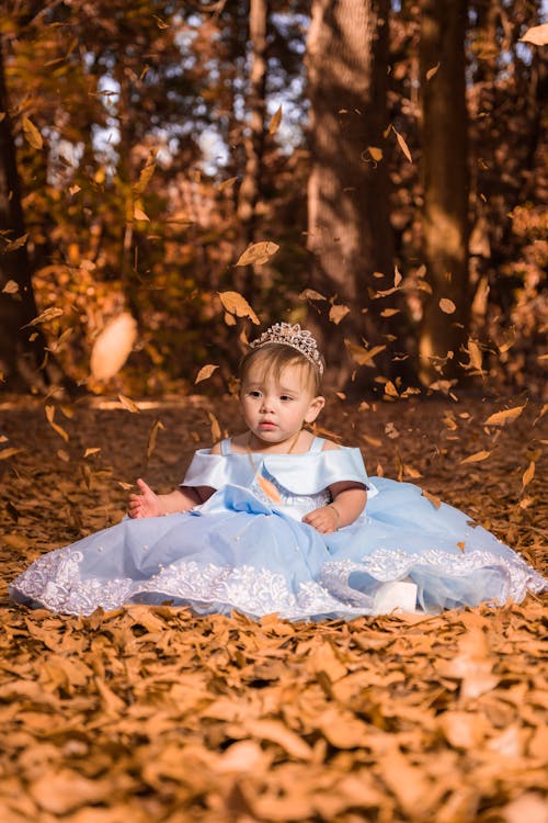 A Cute Baby Girl in Blue Dress Sitting on Dry Leaves