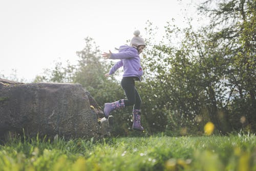 A Girl in Purple Jacket Jumping on a Grassy Field