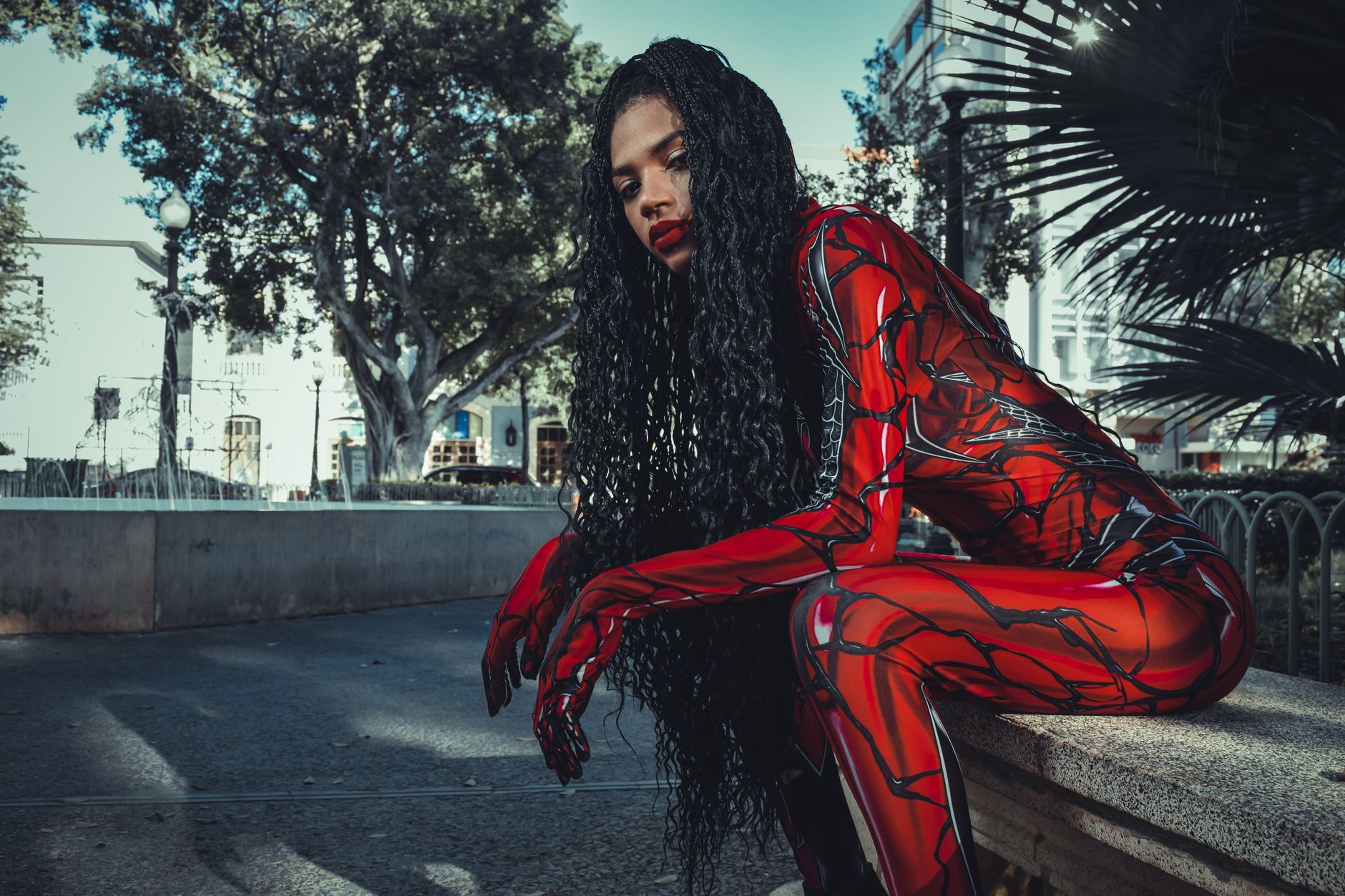 A cosplayer in a vibrant red costume sitting and posing outdoors amidst city buildings and trees.
