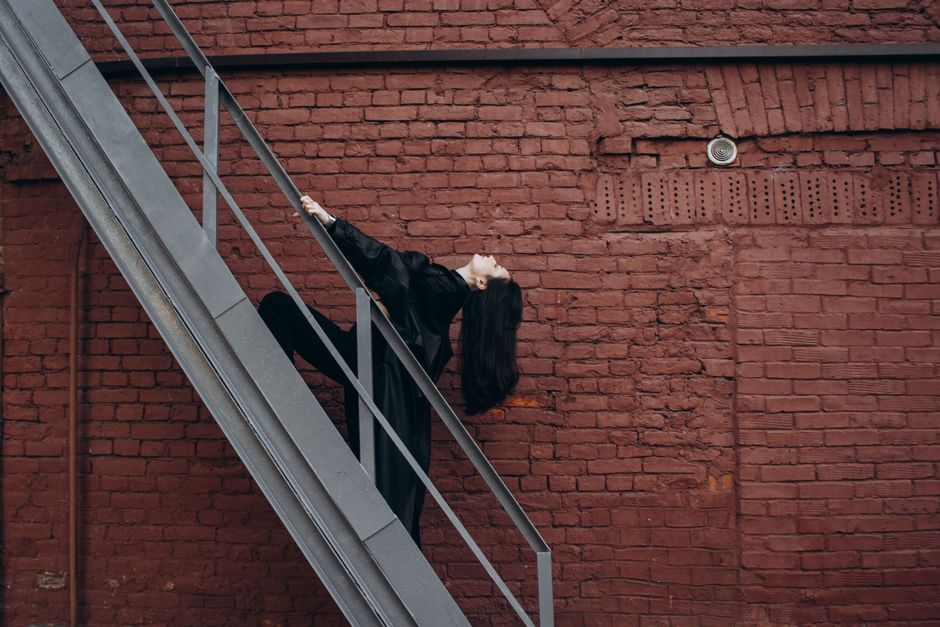Artistic portrait of a woman leaning on an outdoor metal staircase with a brick wall backdrop.