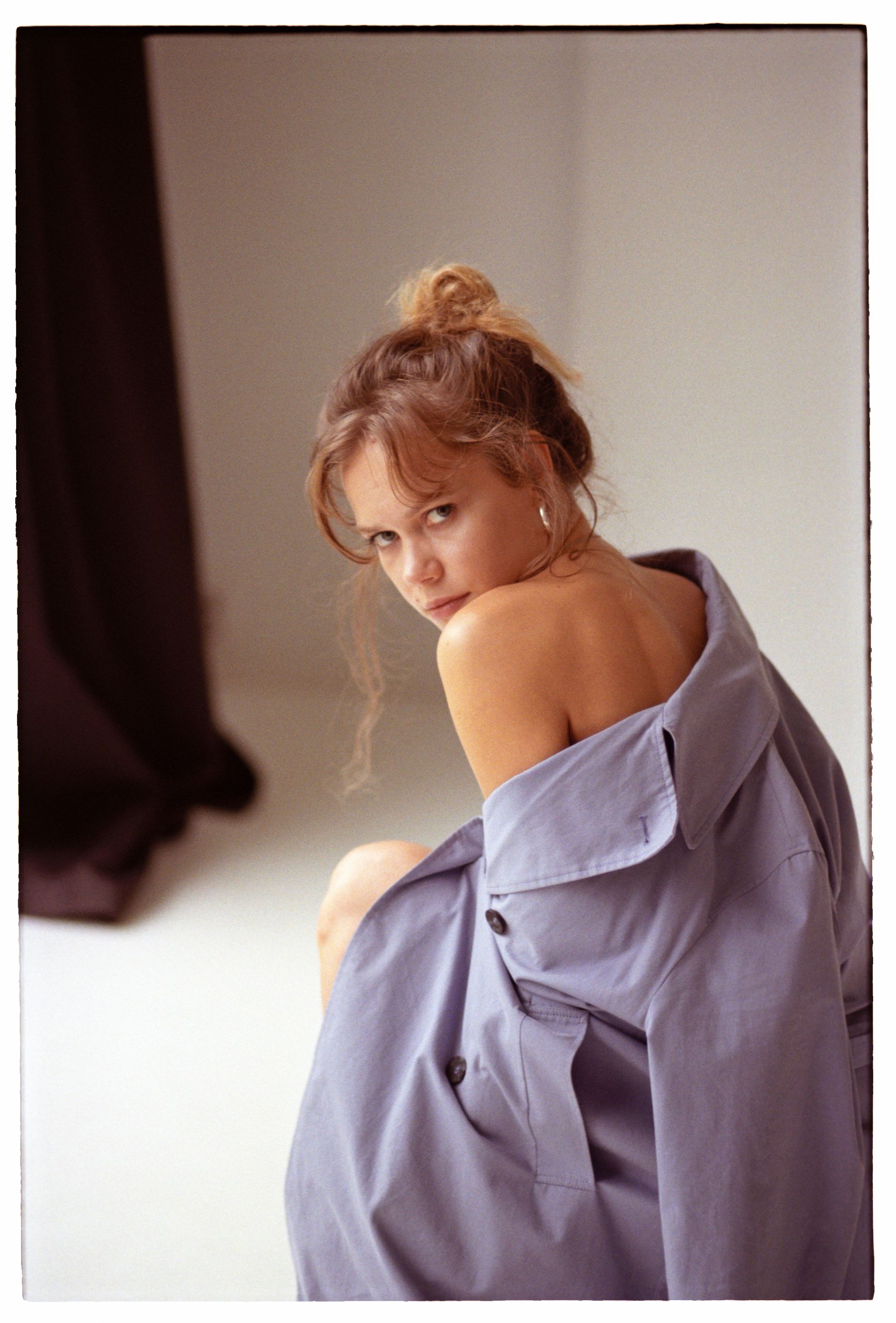 A woman posed indoors wearing an oversized shirt with exposed shoulder, looking back.