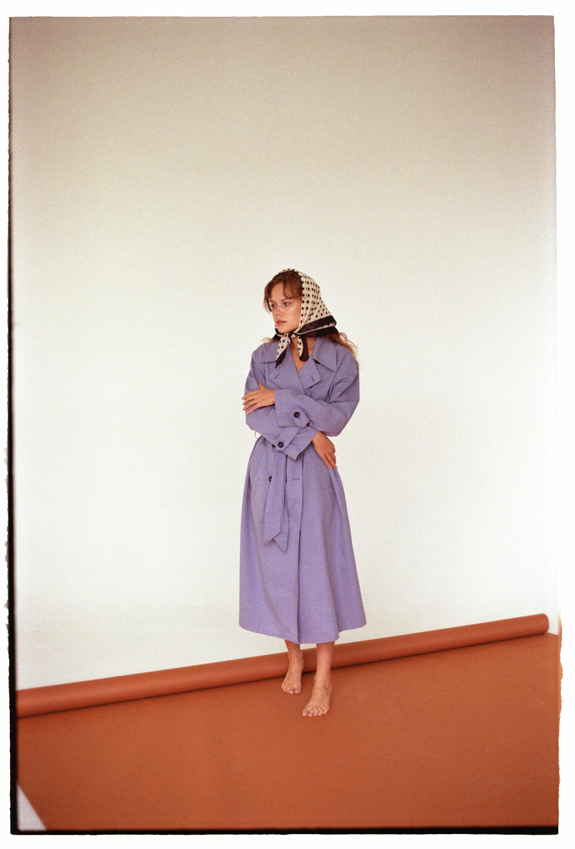 Stylish woman in a purple dress and scarf, posing barefoot indoors, exuding elegance and fashion.