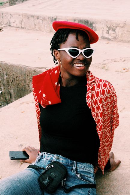 Young black woman in fashionable attire with red beret, sitting and smiling outdoors.
