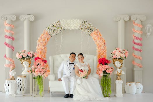 A joyful couple in wedding attire seated under a beautiful floral arch indoors.