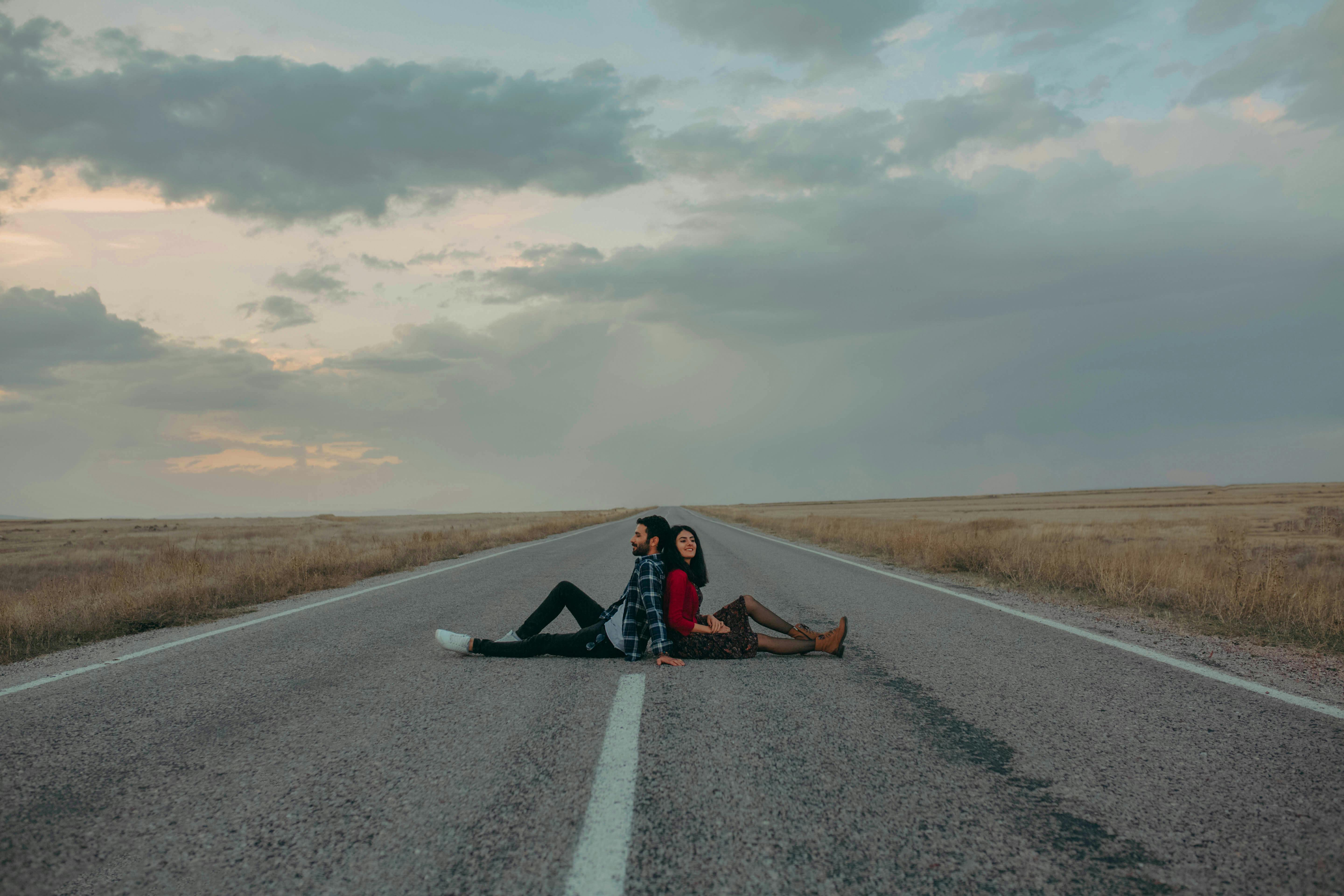 A couple sits back to back on an empty highway under a cloudy sky, evoking adventure.