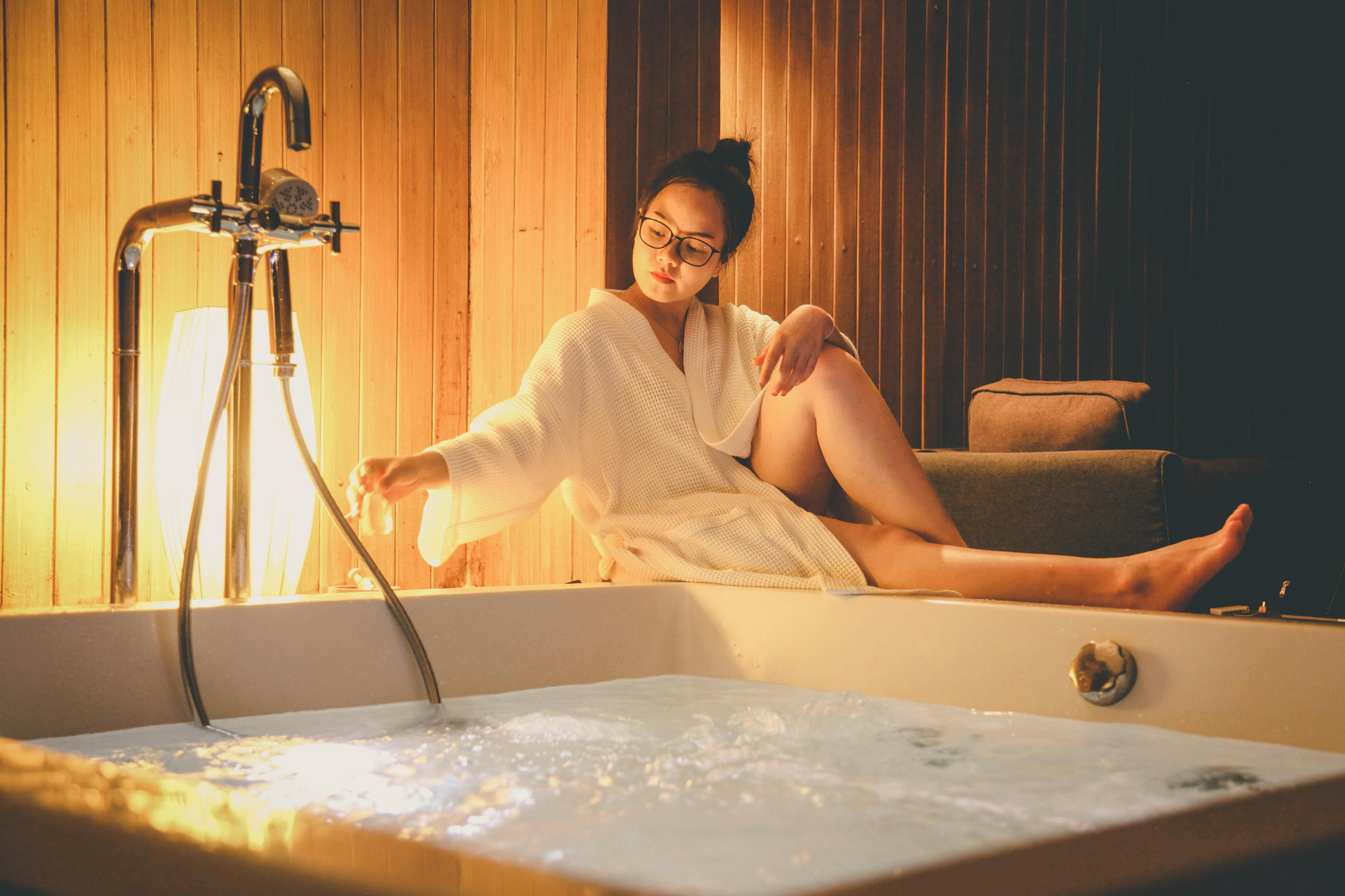 woman wearing white bathrobe sitting beside white bathtub filled with water