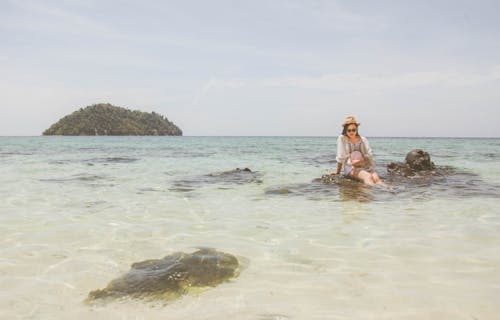 Woman Wearing White Top Sitting on Brown Rock on Body of Water during