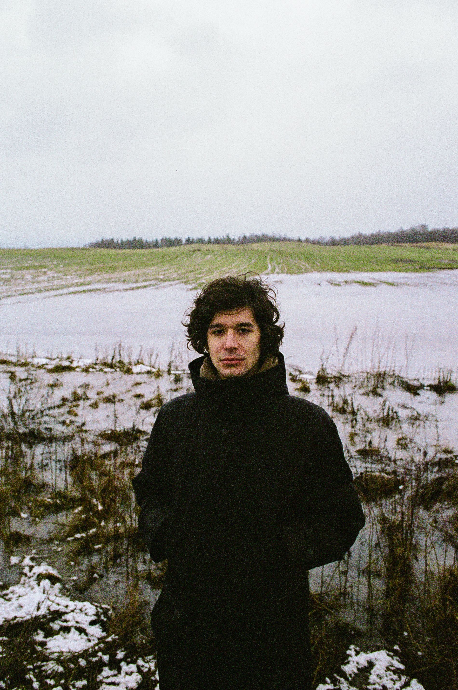 Portrait of a man in a black coat standing in a snowy rural landscape.