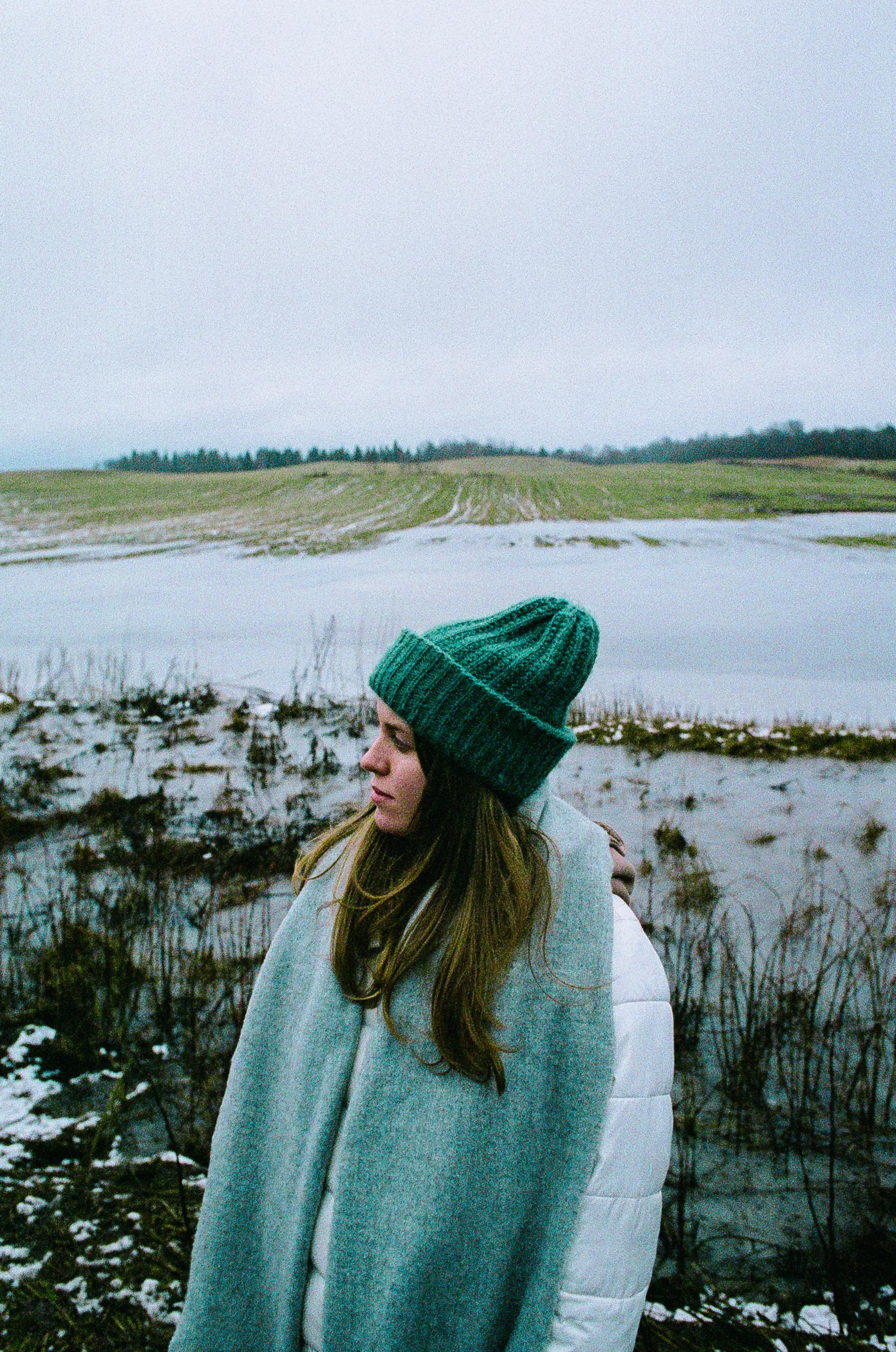 Woman in a green beanie and winter clothing standing in a snowy field with a pond in the background.