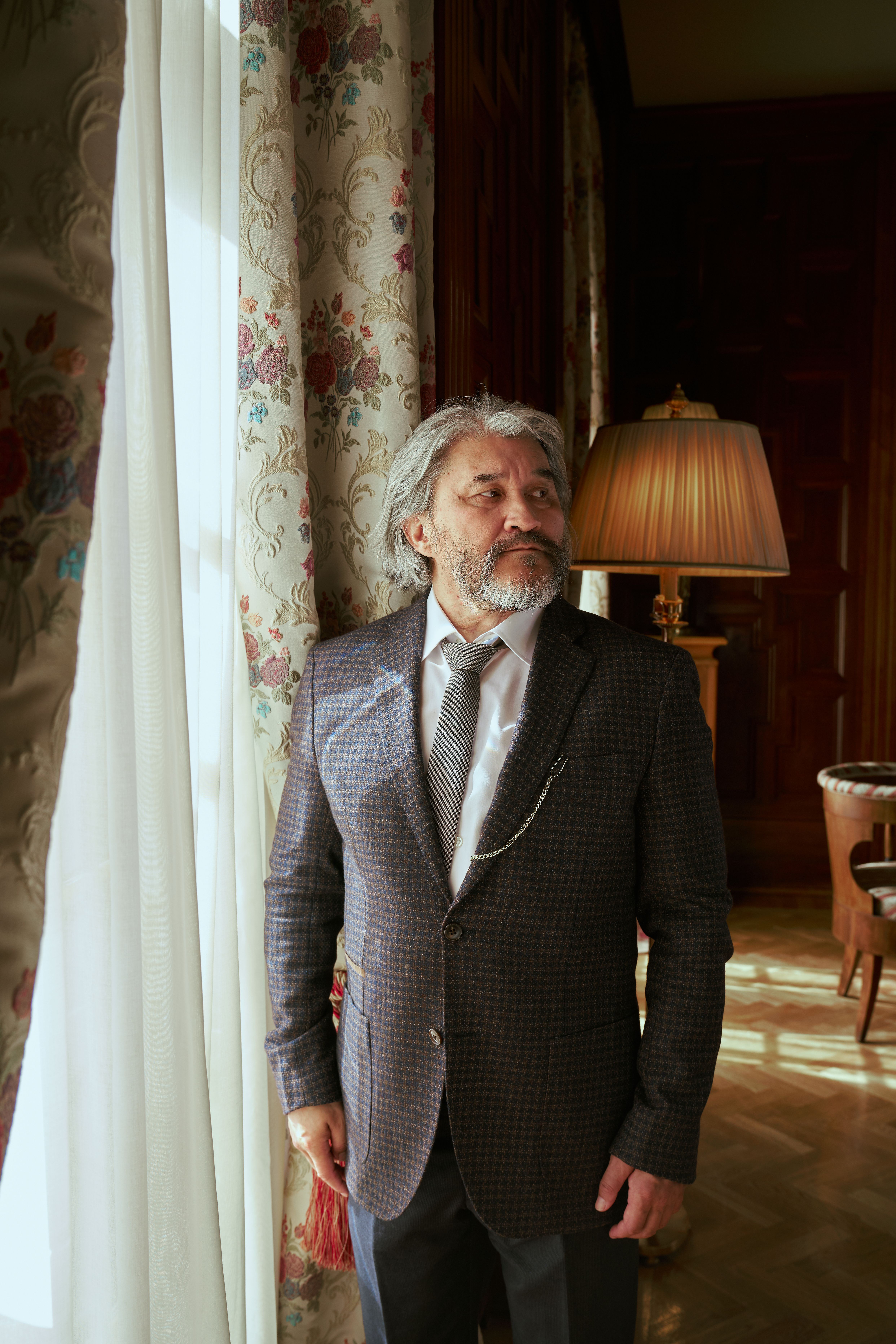 Senior man in a classic suit standing by a window in a luxurious, vintage-style room.