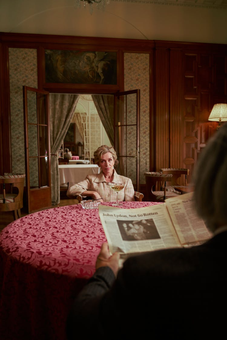 Elegant Woman With Drink Sitting At Table With Man Reading Newspaper