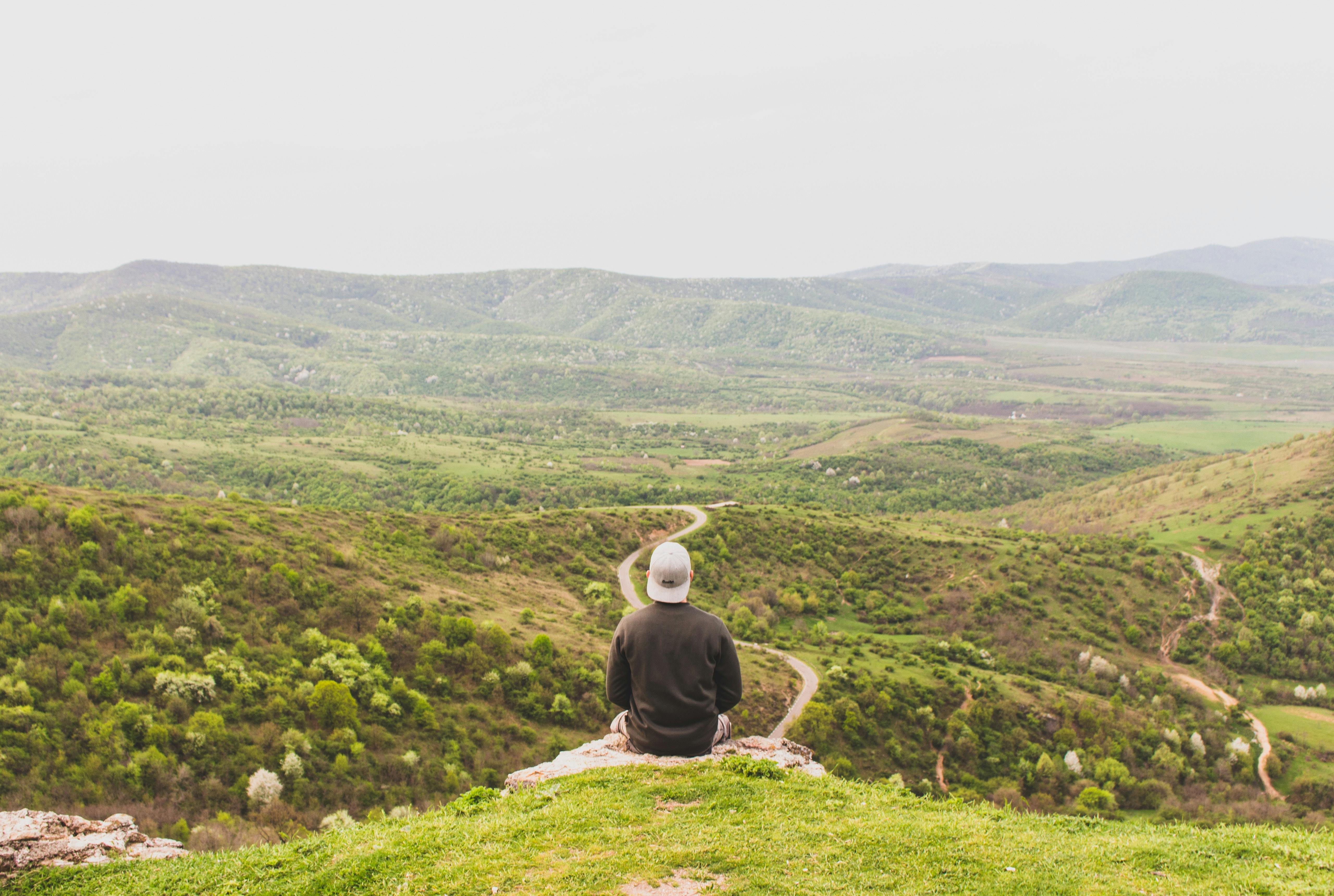 A person sits on a hill overlooking a vast, green valley under a clear sky.