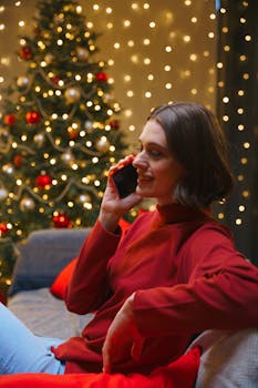 Smiling woman in a red sweater talking on the phone beside a decorated Christmas tree indoors.