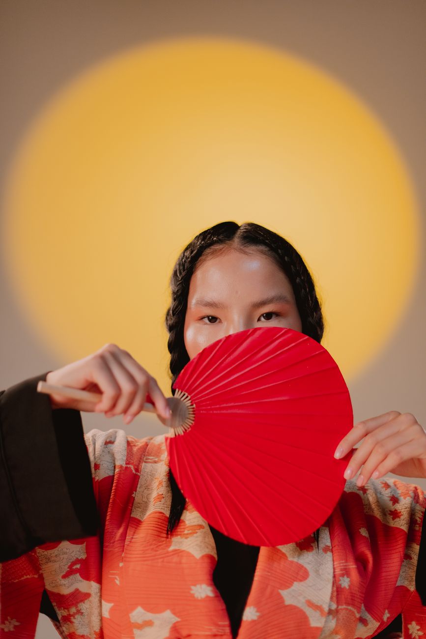 Studio shot of a young woman holding a red hand fan against a warm yellow backdrop.