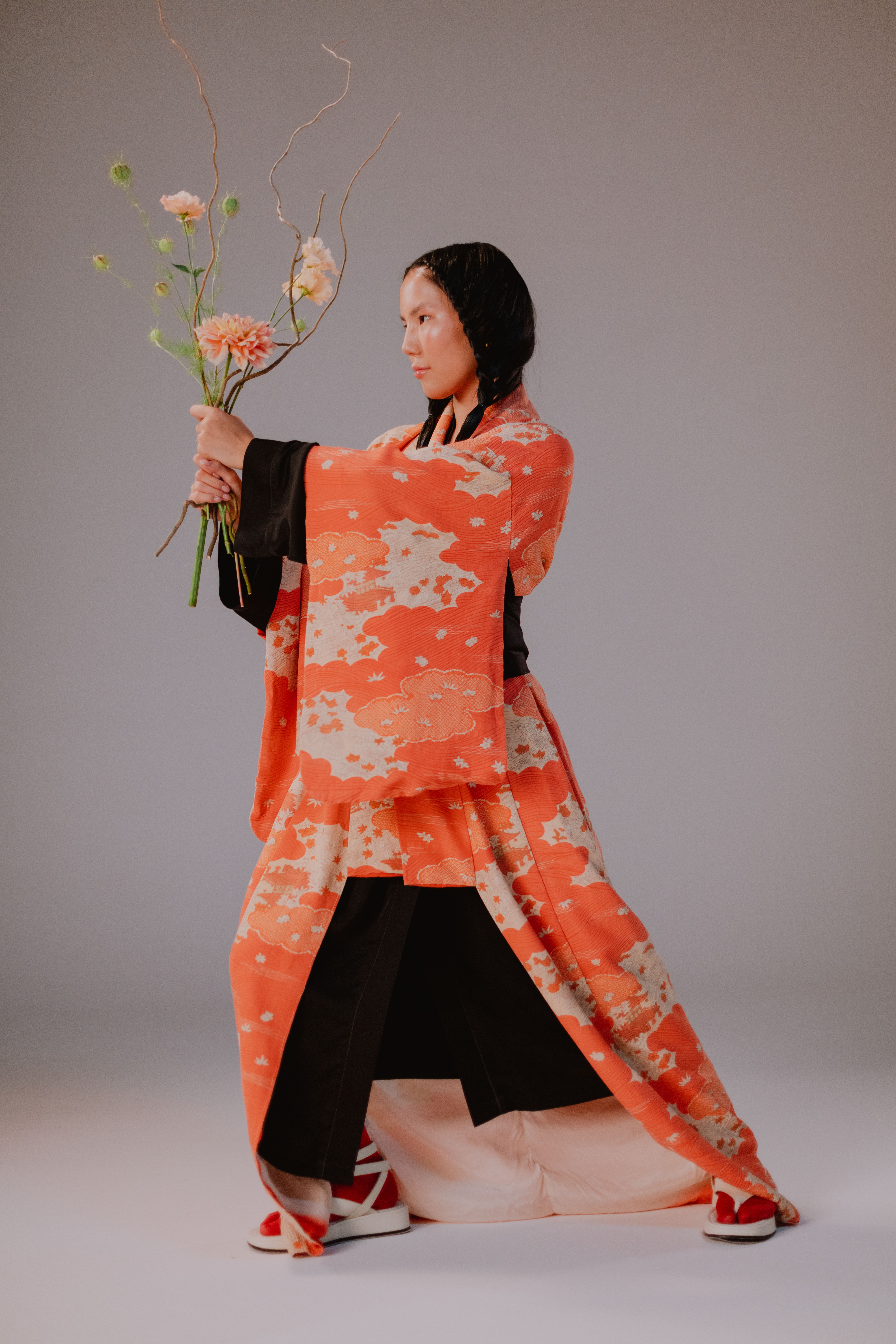 Asian woman in vibrant orange kimono holding flowers, striking a traditional pose.