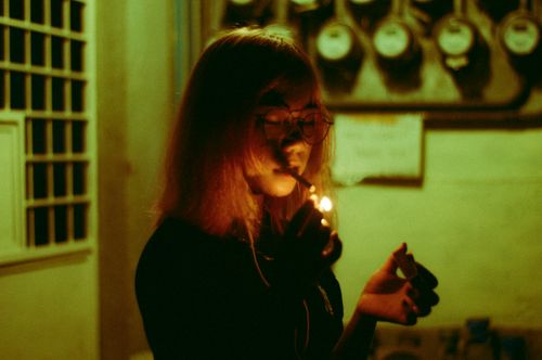 Free A contemplative woman with eyeglasses lights a cigarette outdoors, captured in a moody, dimly lit setting. Stock Photo