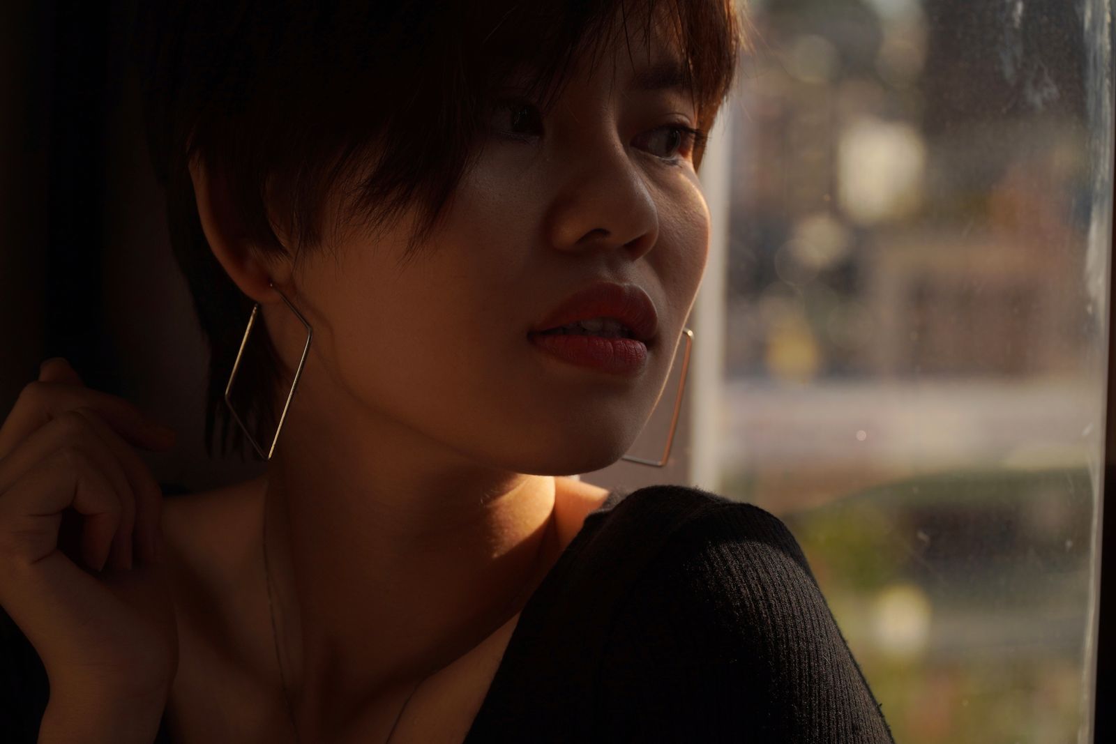 Woman with short dark hair and geometric hoop earrings gazing out a sunlit window
