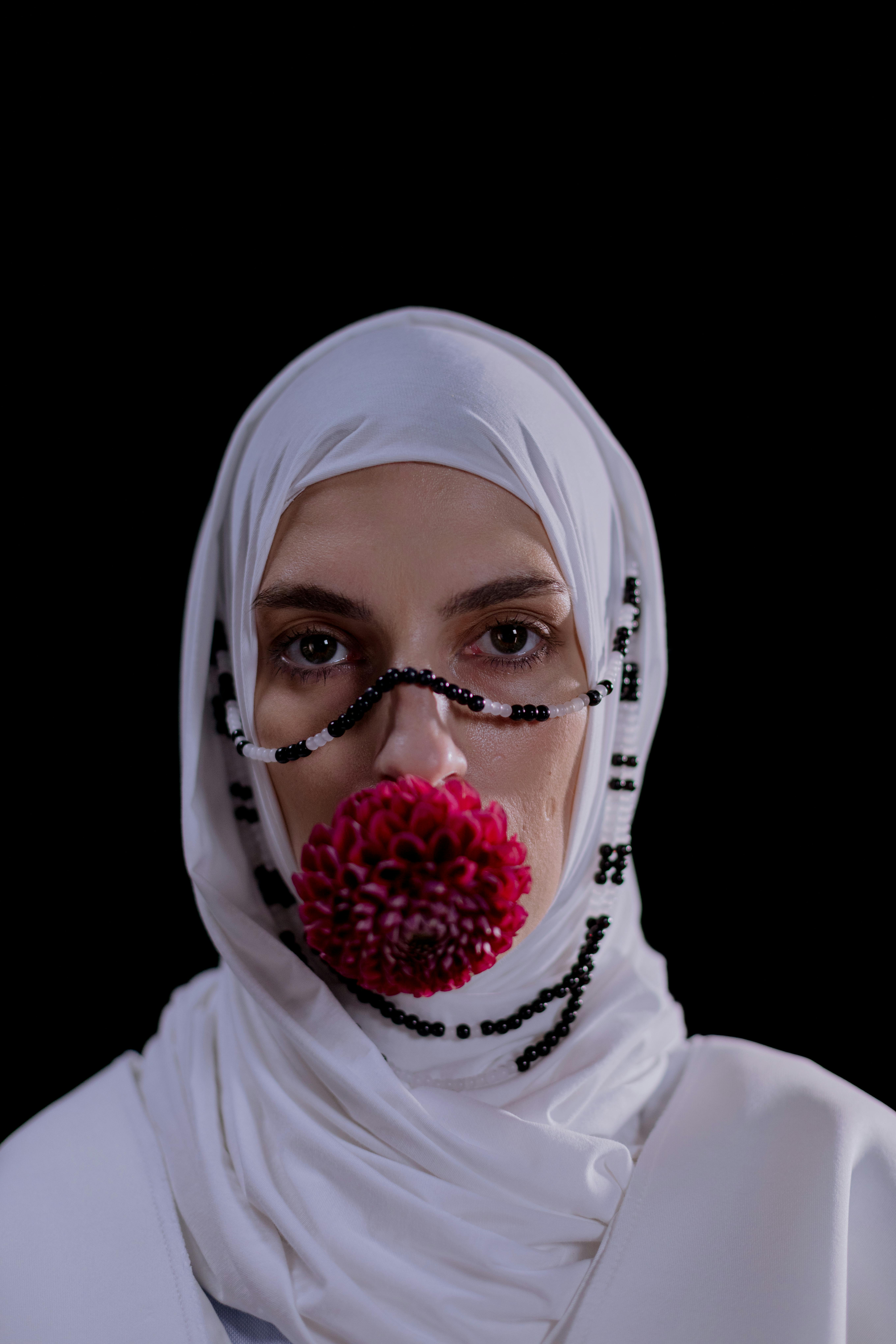 Conceptual portrait of a woman in a white hijab with a flower and bead adornment against a black background.