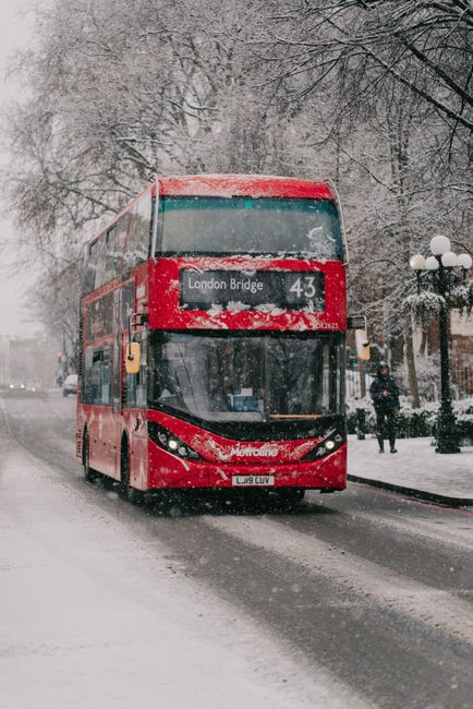 A red double-decker bus drives through snow on a London street in winter.