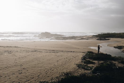 Serene beach scene with a solitary person enjoying the ocean view under a cloudy sky.