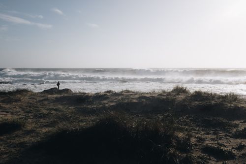 Free A lone person stands on a rugged beach as waves crash onto the shore under a soft early morning light. Stock Photo