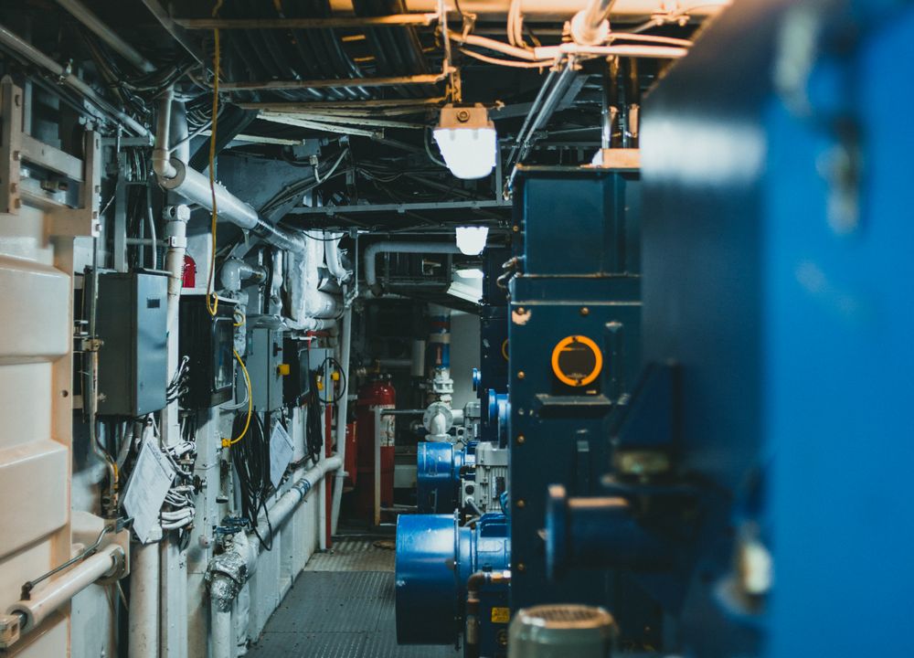 Narrow plant corridor lined with machinery, pipework and valves under service lighting.