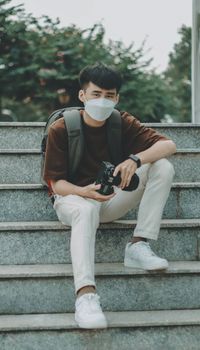 Man wearing face mask sitting on stairs holding a camera, outdoors.