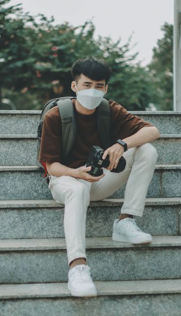 Man wearing face mask sitting on stairs holding a camera, outdoors.