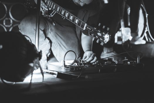 A musician adjusts guitar effects pedals on stage, captured in a candid black and white shot.