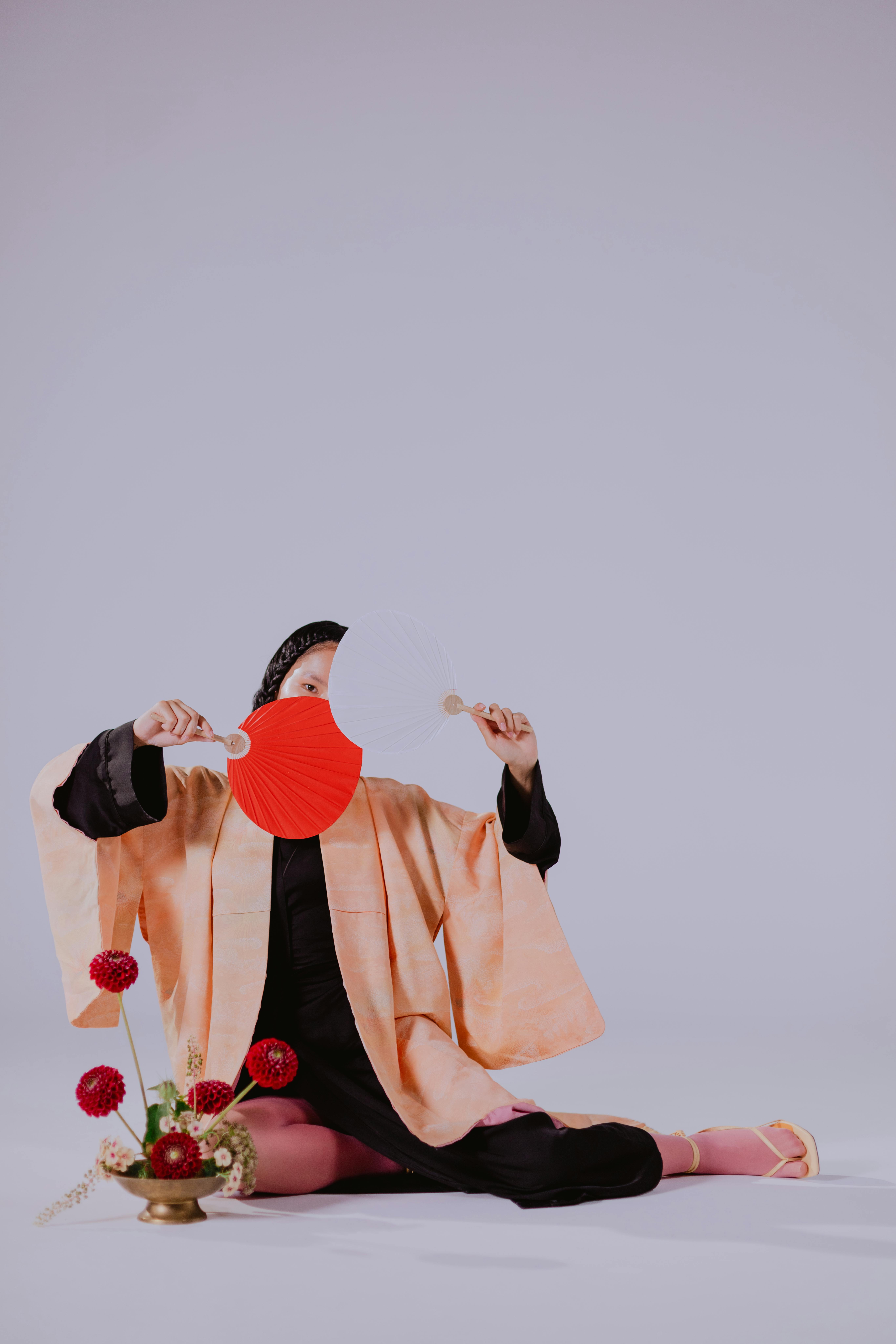 a woman wearing kimono sitting on the floor while covering her face using the hand fans she is holding