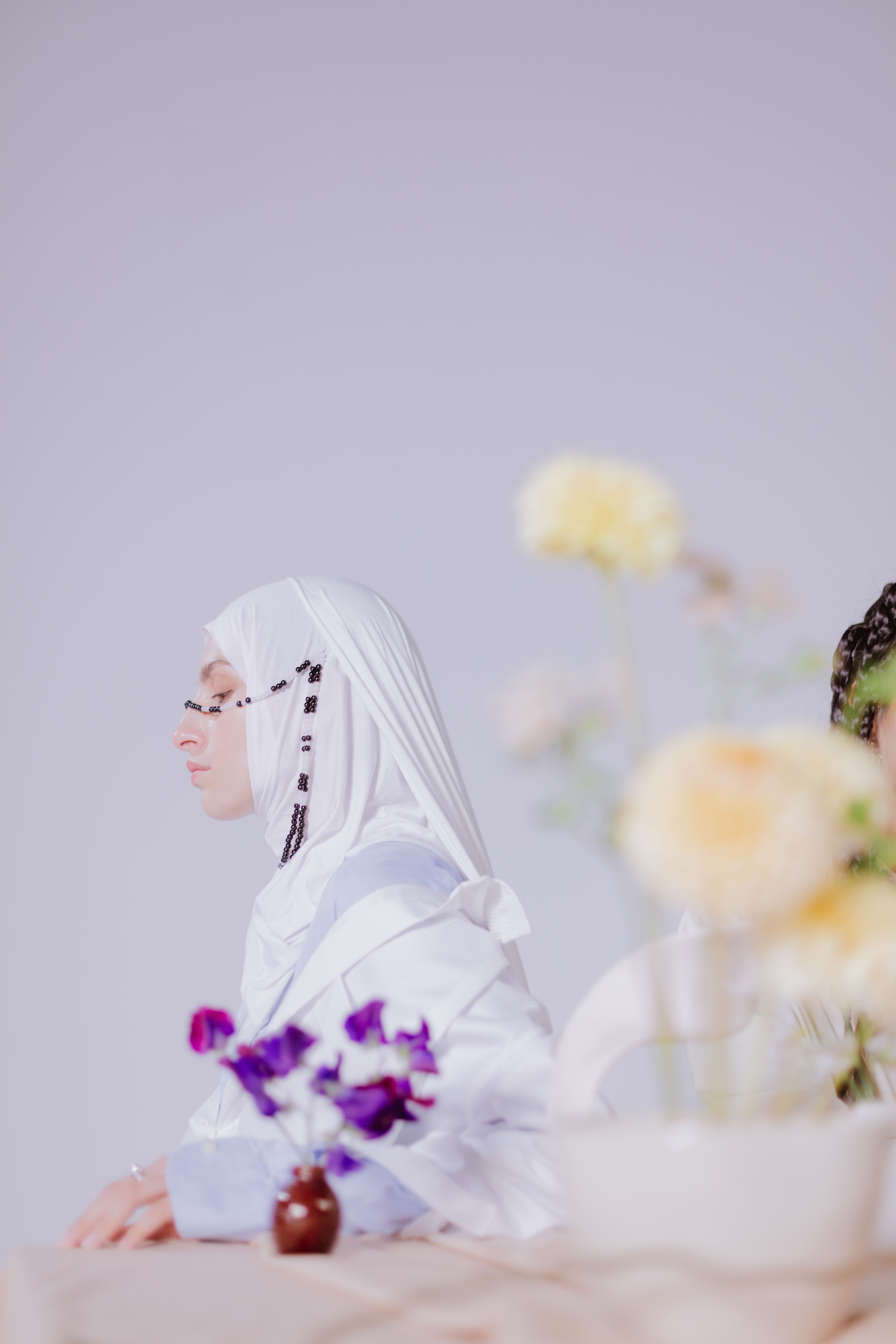 Woman in a white hijab adorned with beads, seated beside delicate flowers on a pastel backdrop.