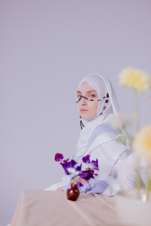 Woman in White Hijab Near Purple Flowers in a Ceramic Vase