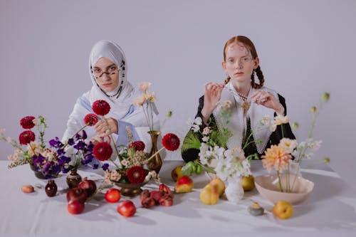 Two Women in Traditional Clothing Sitting at a Table With Fresh Flowers and Fruits