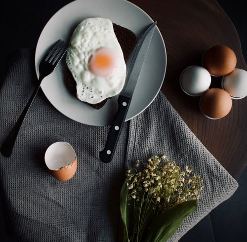 Free Overhead view of a sunny side up egg on toast with cutlery and flowers on a textured cloth. Rustic morning scene. Stock Photo