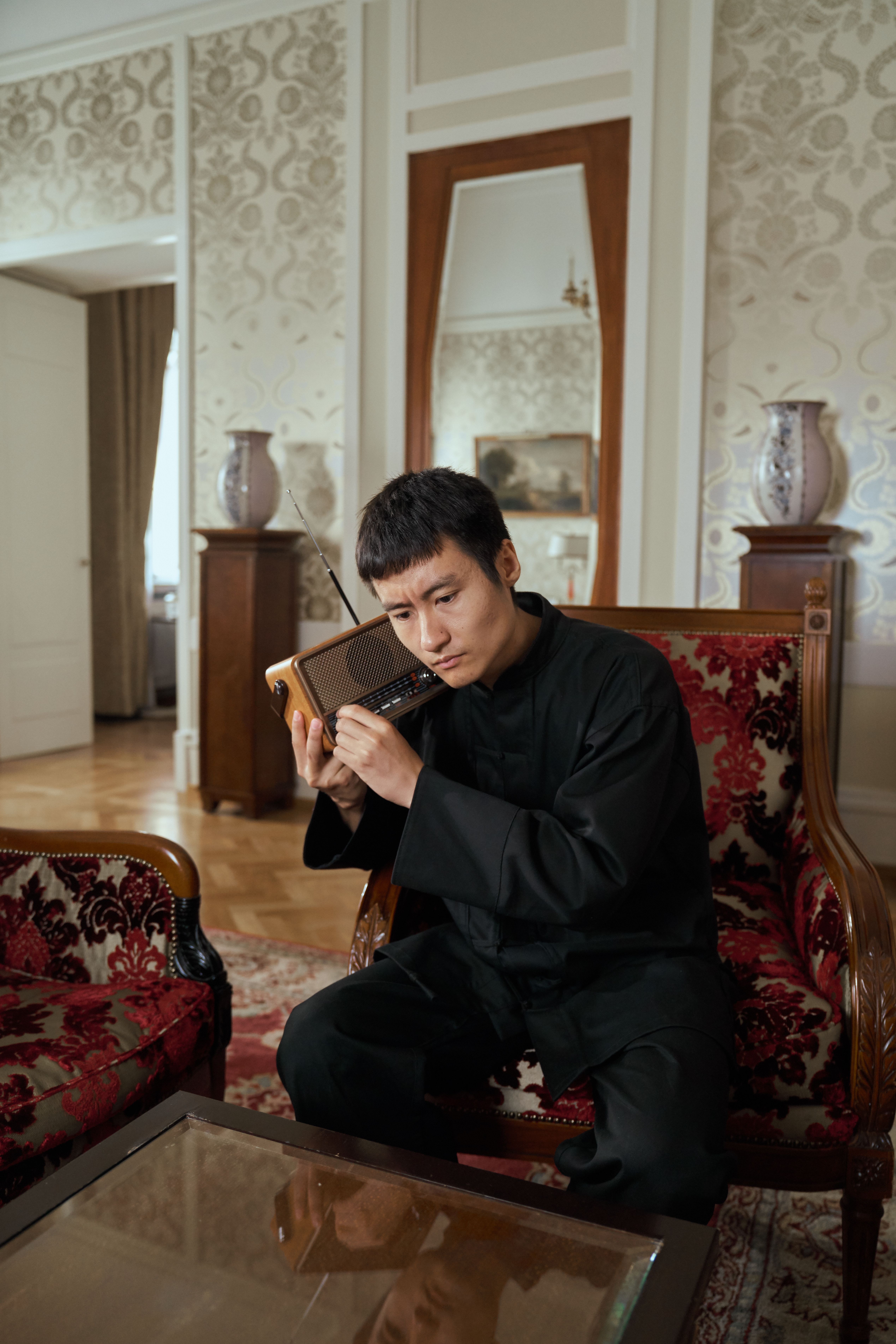 An adult man in traditional attire listens intently to a transistor radio in a vintage-style living room.