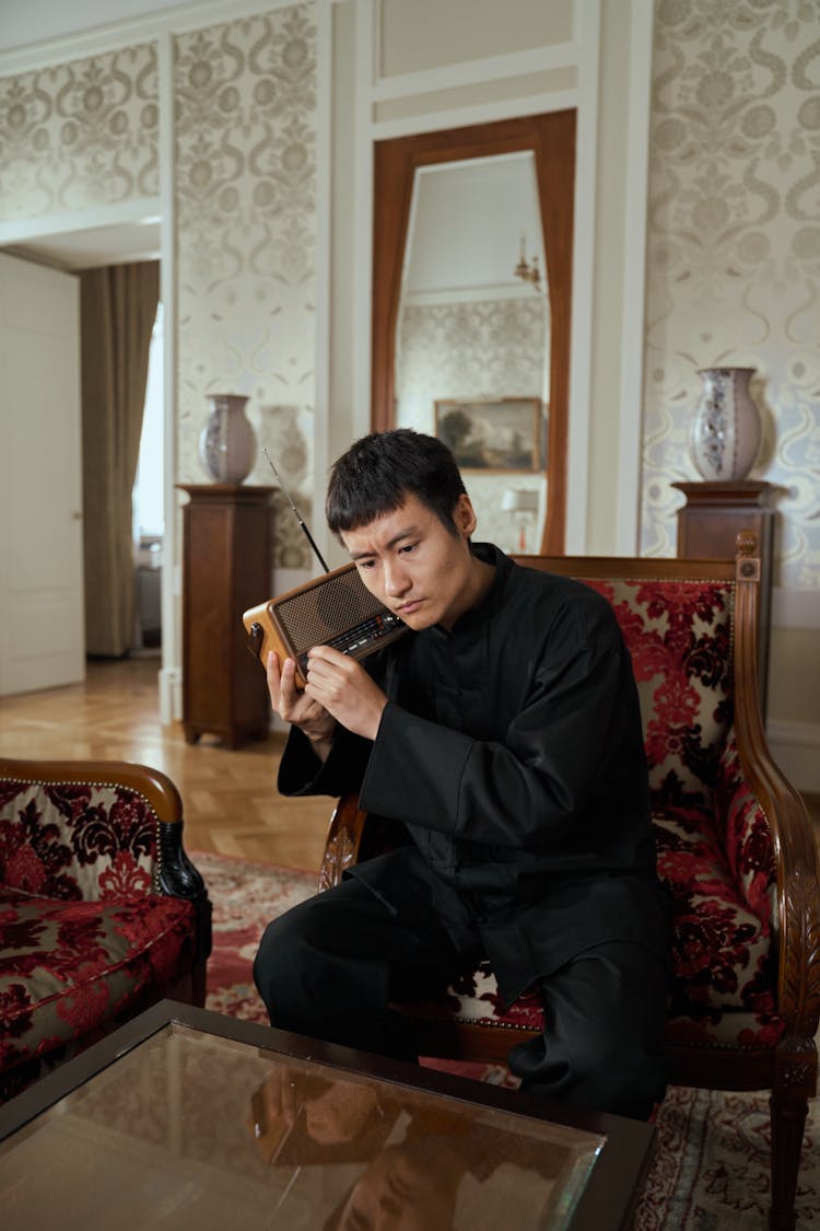 Asian Adult Man Sitting In Retro Armchair And Listening To Transistor Radio