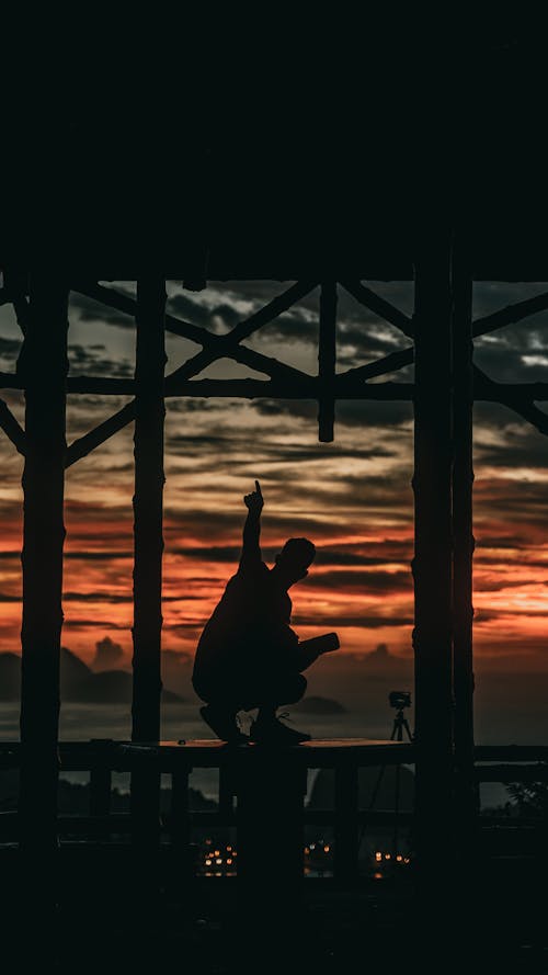 Free A Silhouette of a Person Sitting on a Railing with a Sunset Sky Background Stock Photo