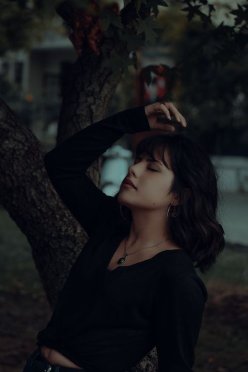 Free A serene young woman with eyes closed enjoys a peaceful moment outdoors leaned against a tree. Stock Photo