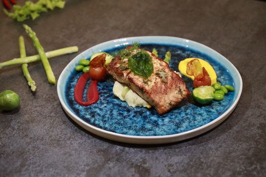 A gourmet salmon dish artfully presented with a variety of vegetables on a blue ceramic plate.